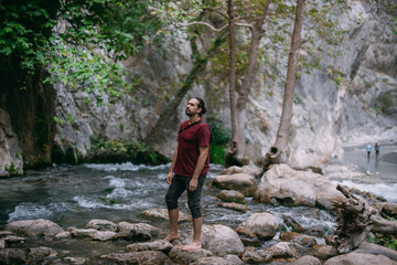 Naklejka na ściany i meble A young male tourist near a clear mountain stream in the rocks overgrown with greenery, on a summer day