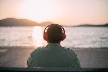 A young man is sitting on the beach with headphones on at sunset by the sea.