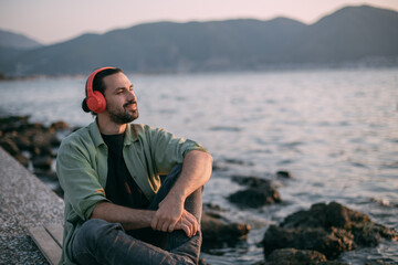 A young man is sitting on the beach with headphones on at sunset by the sea.