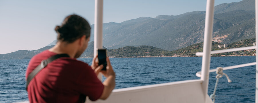 A young, contented man, a tourist rides a boat to the sea, takes pictures on the phone, enjoys a sunny day.