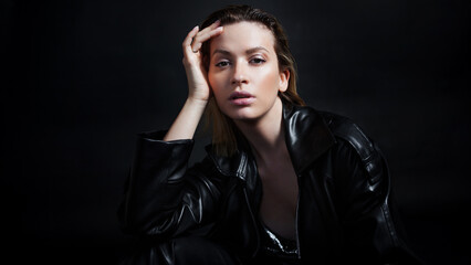 Stylish portrait of a young beautiful woman in a black leather raincoat on a black background. Trendy beautiful blonde