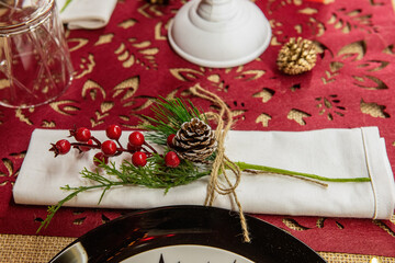 Festive Christmas table setting. On the red runner lies a white napkin, decorated with a branch of holly, with a snow-covered cone, tied with a string. New year home decor. Copy space
