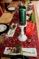 Festive Christmas table setting. On a wooden table lies a red runner, black plates on white says Enjoy, golden forks, spoons, knives. New year green candles, fir branches with cone, Holly. Style decor