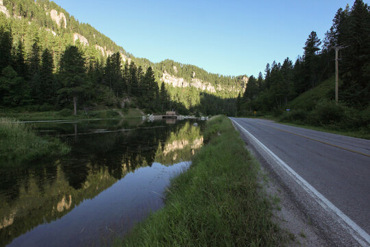 A stream runs parallel open road at the base of a mountain valley.; Deadwood, South Dakota
