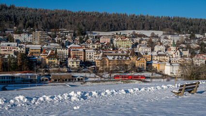 Winter landscape with snow. Red train crossing Tramelan village in sunny day. Bern Canton,...