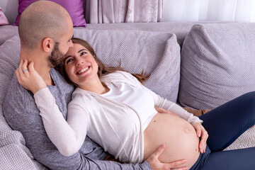 A happy loving couple resting on the sofa, preparing for childbirth. Pregnant woman embraced around her big tummy by her partner.