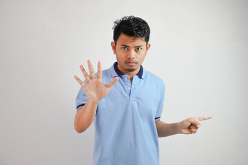 Attractive Asian man with hand gesture pose rejection or prohibition while pointing to copy space by his side wearing blue t shirt isolated on white background