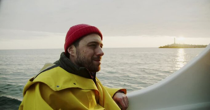 A sailor sits on a boat and smilingly looks at a lighthouse in the distance