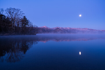 妖しいグラデーションの空の月と山々を湖面に映す朝靄の漂う夜明けの湖。