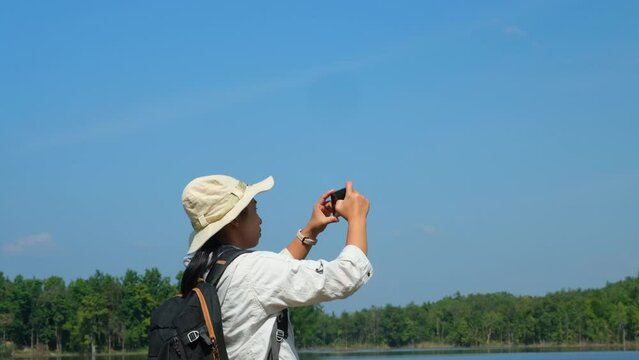 Female ecologist with backpack using mobile phone taking picture of reservoir for data analysis. Female environmental scientist working in field survey.