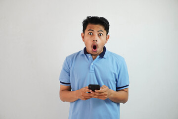 shocked young asian man holding phone and open mouth wearing blue t shirt isolated on white background