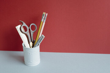Ruler, scissors, colored pencil, pen in pencil holder case on gray desk. red wall background. copy...