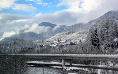 A bridge over a mountain river and a village covered with snow on the background of mountains in fog