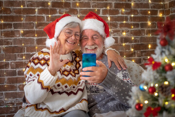 Smiling senior adult couple sitting at home at Christmas time in video call with family or friends. Lovely elderly man and woman stay together at celebration event