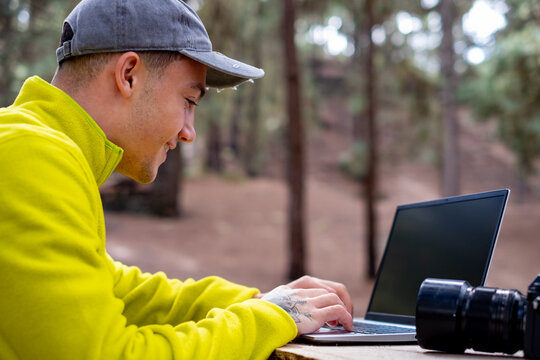 Business on fresh air. Handsome smiling young protographer man working on laptop while sitting at the wooden table outdoors in the forest. Concept of smart working in nature