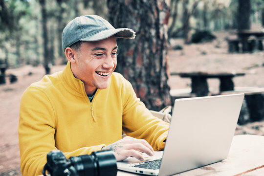 Business on fresh air. Handsome happy young protographer man working on laptop while sitting at the wooden table outdoors in the forest. Concept of smart working in nature