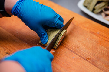 Hands in blue gloves cut a snack roll with greens, cheese and fish.