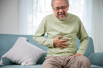 Fototapeta premium Elderly Asian old man sitting on sofa having stomachache, old age suffering from stomach ache holding his stomach in living room, people health problem, food poisoning