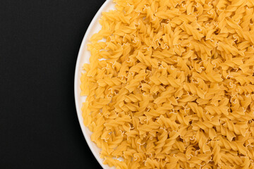 Uncooked Fusilli Pasta Lying on White Plate on Black Background. Raw and Dry Macaroni. Unhealthy and Fat Food - Top View