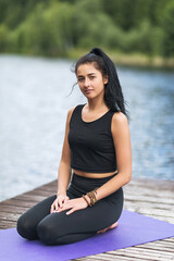 Fototapeta na wymiar Portrait of a young beautiful female yoga trainer sitting on a wooden footbridge on the shore of a lake