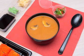 Delicious Japanese tom yum soup, rolls with salmon, avocado, cucumber, cheese in takeout packaging with wood sticks and soy sauce. Selective focus. Asian food and delivery concept. Healthy eating