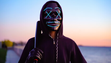 Mysterious Halloween mask. Man in the blue mask glow with a bat in hand at dusk at sunset by the...