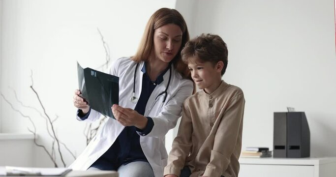 Young female pediatrician or traumatologist holding roentgen film, showing x-ray with radiology results to little curious boy, explaining treatment after trauma to small patient. Pediatry, childcare