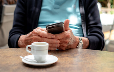 Fototapeta na wymiar Closeup on old senior man hands using mobile phone. Elderly caucasian male sitting at cafe table with an espresso coffee cup while looking at his smartphone