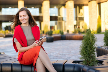 Close-up portrait of a charming young woman in a red skirt sitting on the street with a phone in her hands. Smiling and looking into the camera, head turned to the side
