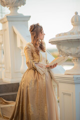 A beautiful young woman in a historic eighteenth century gold dress stands on the stairs of the...
