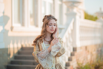 A beautiful young woman in a historic eighteenth century gold dress stands on the stairs of the...