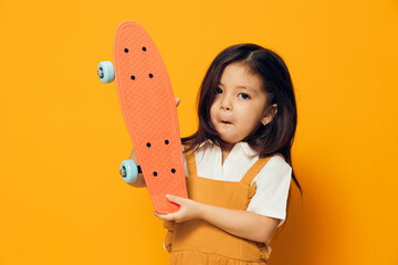 funny, cheerful, emotional little girl in a sundress stands holding a small skateboard in her hand...