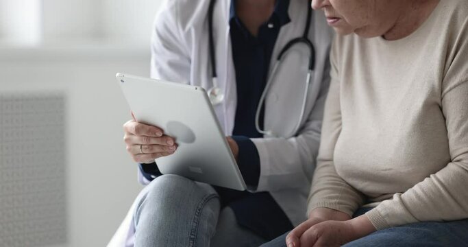 Cropped close up view unknown elderly patient listen to woman doctor during visit in clinic, gp teach older female to use new medical application, show health test results on tablet device. Healthcare