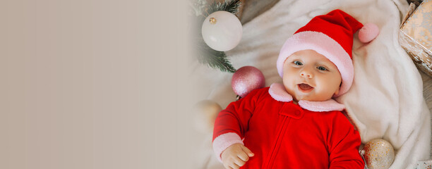 Close-up portrait of a newborn baby. Cute Caucasian baby 5-6 months old in Santa costume lying on a cozy blanket near a decorated Christmas tree. Merry Christmas 2022