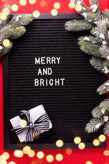 Felt or letter board with phrase merry and bright, box with present and fir tree spruces on bright red paper textured background. Top view. Bokeh light. - 554425705
