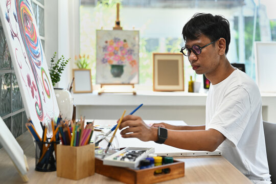 Asian man artist sitting in art studio and painting picture on canvas with oil paints. Art and leisure activity concept