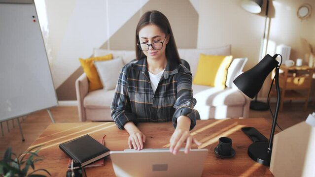 Girl in home office on table, at place of work opens laptop screen and starts working. Young, beautiful and business woman in glasses typing and looking at computer monitor. Online freelance.