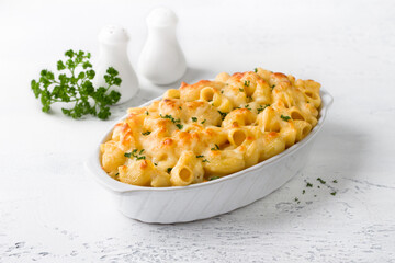 Mac and cheese, baked American pasta in cheese sauce decorated with parsley in a baking dish on a light gray background. Delicious homemade food - 554430986