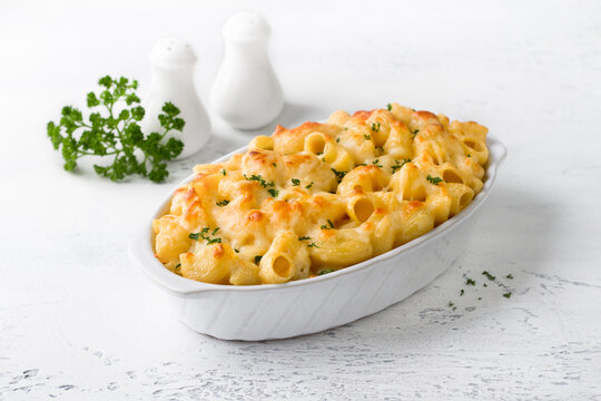 Mac and cheese, baked American pasta in cheese sauce decorated with parsley in a baking dish on a light gray background. Delicious homemade food