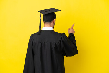 Young university graduate caucasian man isolated on yellow background pointing back with the index...
