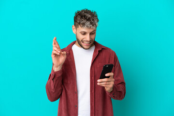 Young handsome caucasian man isolated on blue background using mobile phone with fingers crossing
