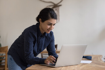 Focused happy young Indian laptop user woman standing at workplace table in stylish home office,...