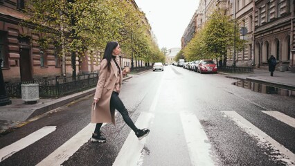 Business woman walks at street of city center and crosses road on zebra, hold digital tablet in hands. Atmospheric autumn outfit with coat. Nature with trees and old beautiful architectural buildings. - Powered by Adobe