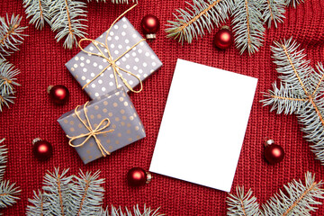 Christmas greeting card mockup, gift box, fir tree branches and holiday decorations on red knitted sweater background, top view, flat lay. White New Year card with winter decor
