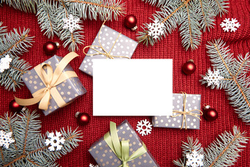 Christmas greeting card mockup, gift box, fir tree branches and holiday decorations on red knitted sweater background, top view, flat lay. White New Year card with winter decor