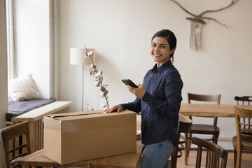 Happy Indian renter girl leaving apartment, packing box for moving, ordering courier, delivery,...