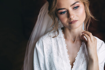 a pensive bride in a white robe and veil straightens the necklace around her neck. preparations in...