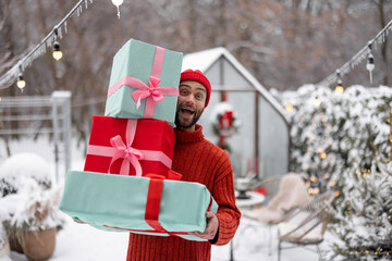 Cheerful man in red sweater and hat carries gift boxes at snowy backyard. Concept of happy winter...
