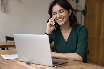 Cheerful employee woman using digital gadgets for online wireless communications in home office, sitting at work table, making business call, talking on mobile phone at laptop, smiling, laughing
