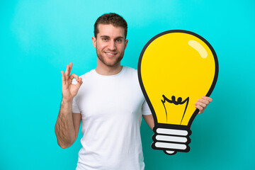 Fototapeta na wymiar Young caucasian man isolated on blue background holding a bulb icon and doing OK sign